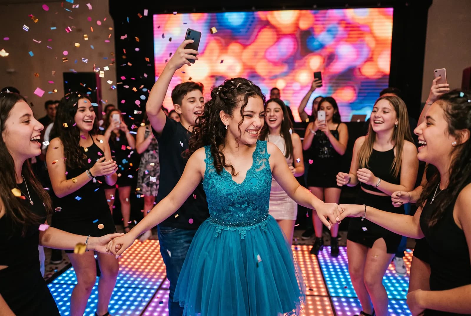 Quinceañera con vestido corto turquesa bailando con sus amigas en una pista iluminada, con una pantalla LED detrás y papelitos en el aire